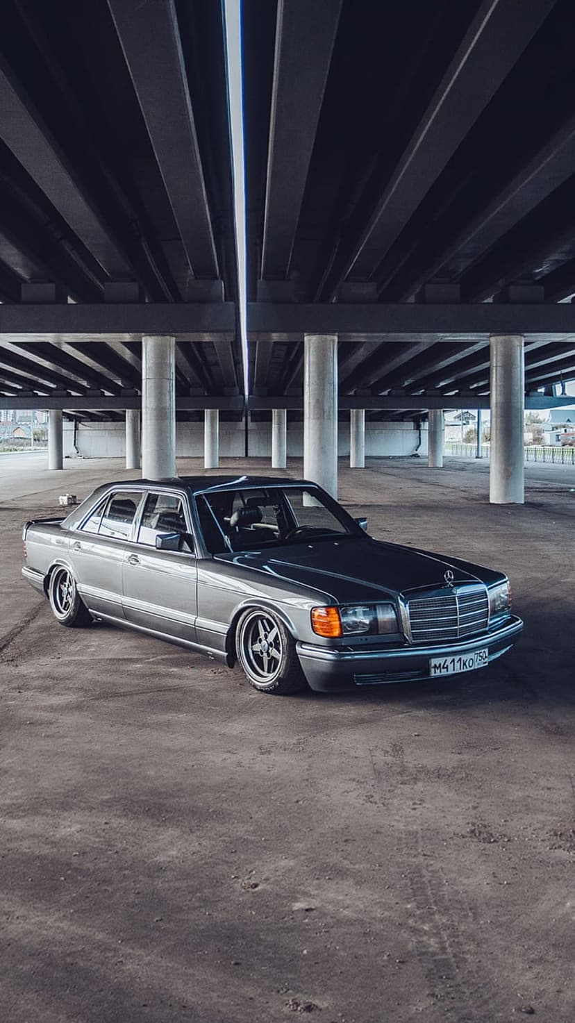 Mercedes-Benz W126 sedan with custom wheels parked on concrete under a massive grey brutalist bridge structure.
