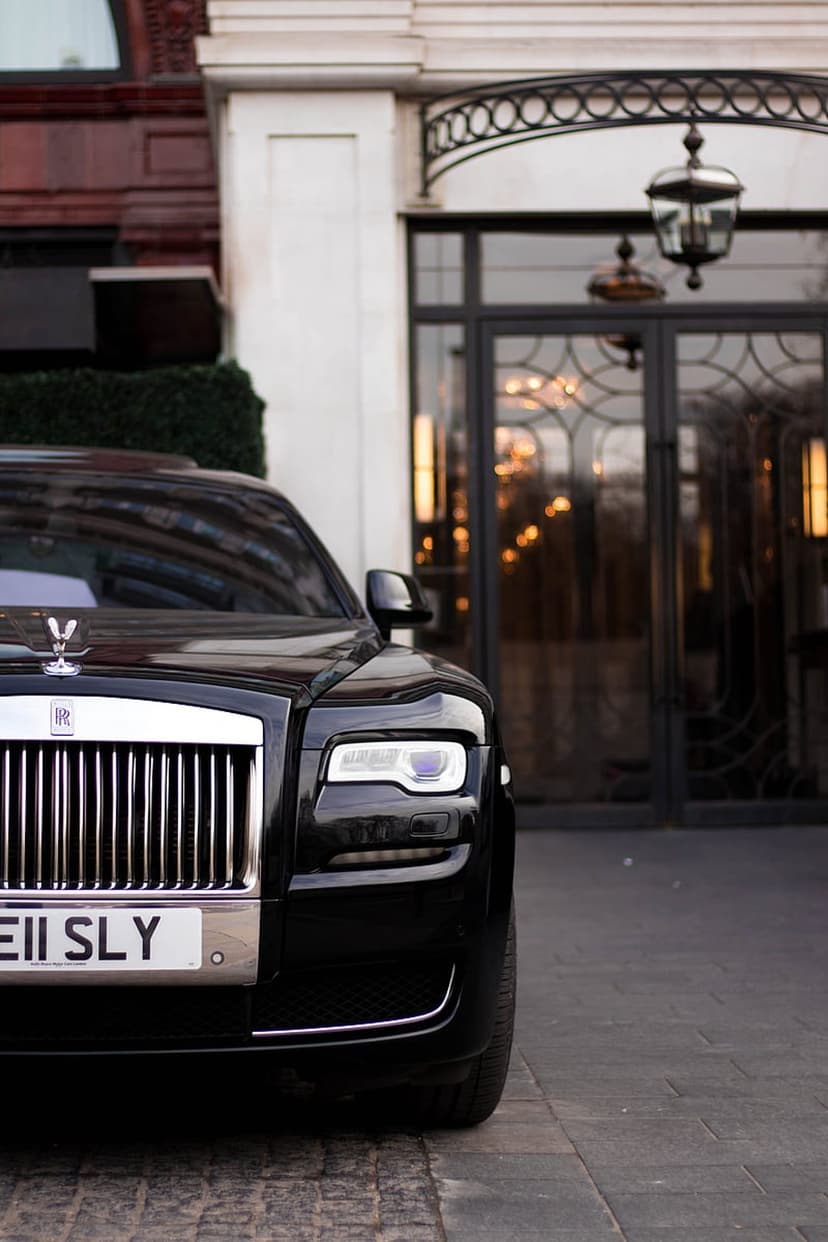 Black Rolls-Royce Ghost with glowing headlights parked on cobblestones in front of a grand building entrance.