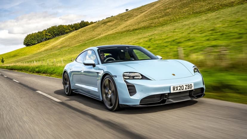 Light blue Porsche Taycan driving on a rural road past green hills with motion blur and bright daylight.