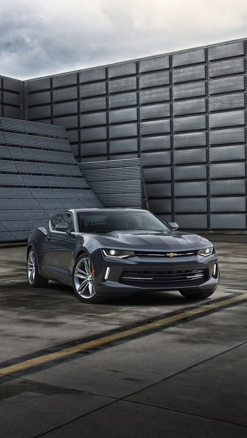 Dark grey Chevrolet Camaro parked on concrete with yellow lines before a layered metallic industrial building.