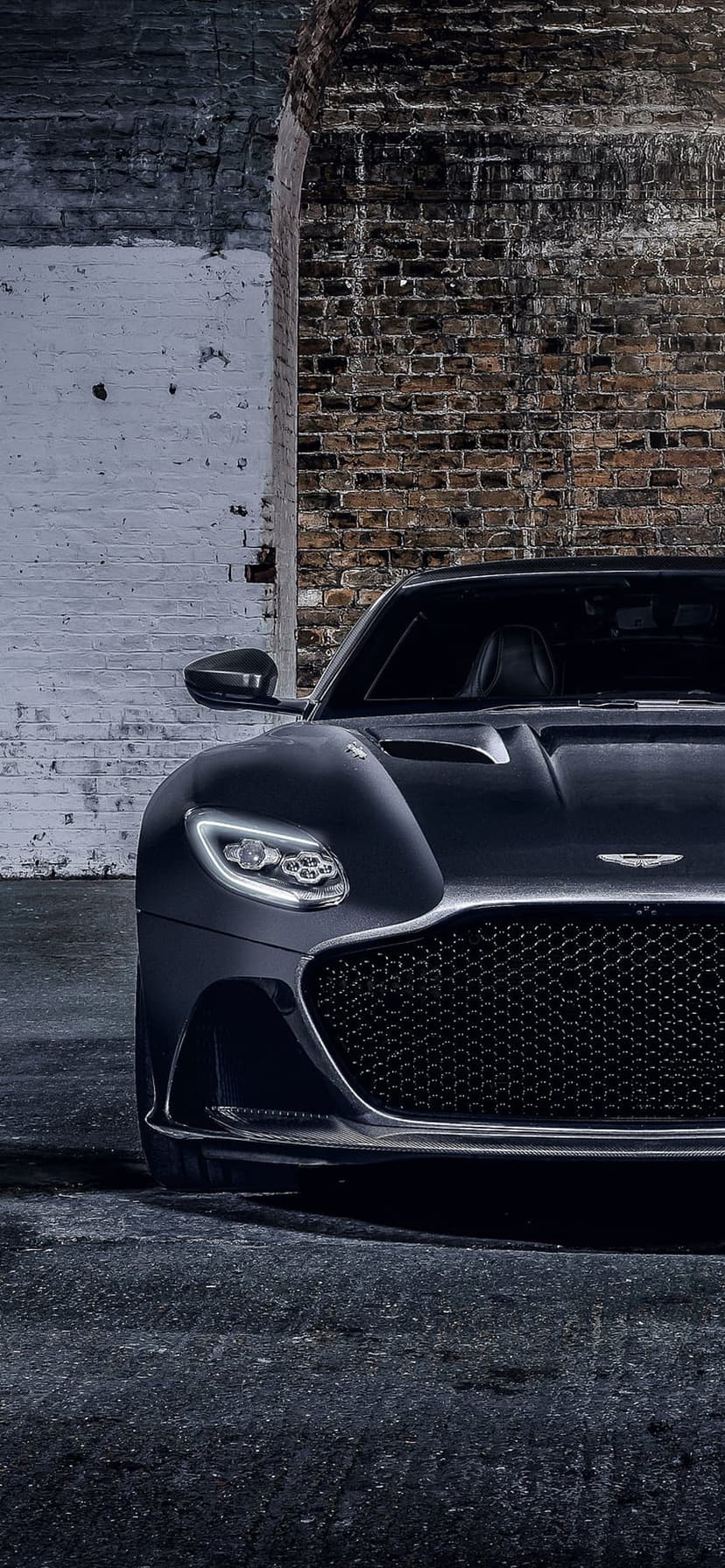 Aston Martin DBS Superleggera with prominent front grille parked on asphalt against a textured brick wall.