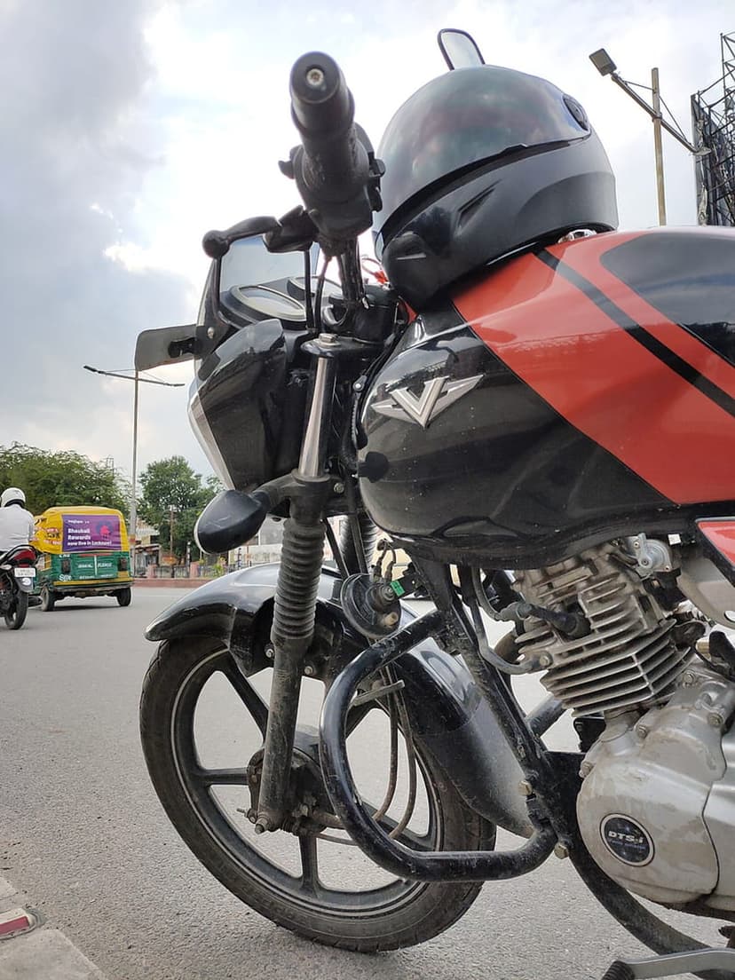 Black motorcycle with a helmet on the handlebars parked near a yellow auto rickshaw on an asphalt city street.