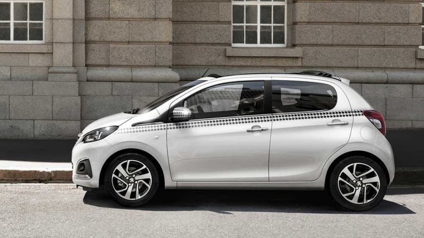 Side view of a white Peugeot 108 city car with a houndstooth decal parked against a textured stone wall.