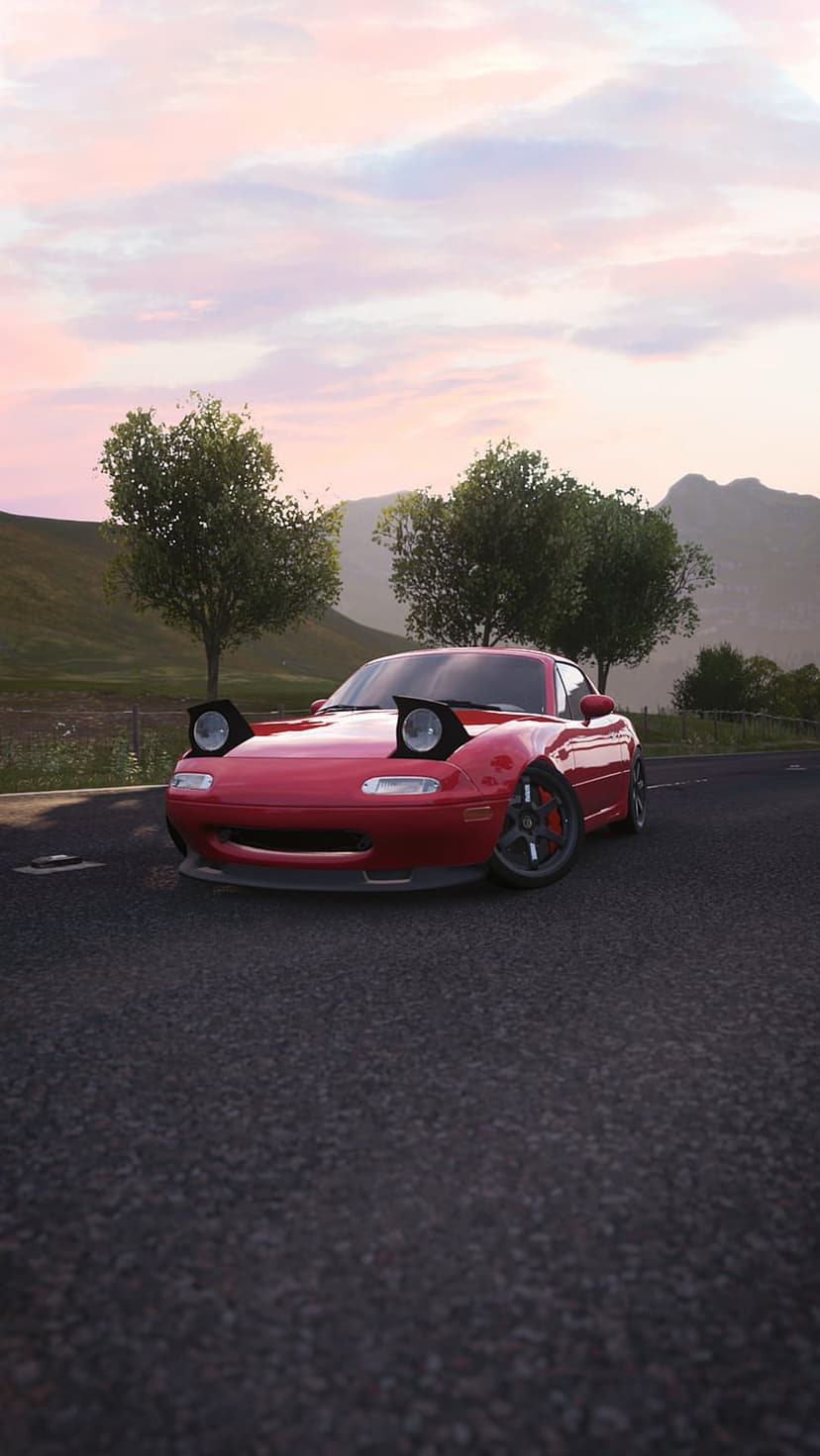 Red Mazda Miata NA with pop-up headlights up on a paved road during a pastel sunset with green hills behind it.