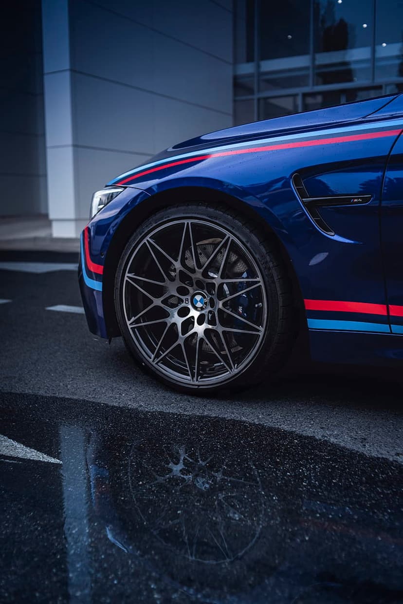 BMW M4 in blue with racing stripes parked on wet pavement reflecting the alloy wheels and chassis.