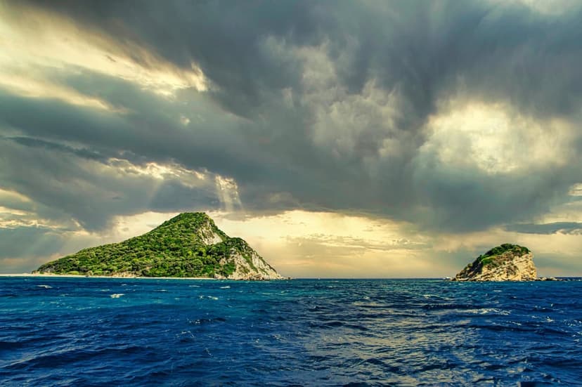 Lush green islands rise from a choppy dark blue ocean beneath heavy storm clouds and dramatic rays of sunlight.