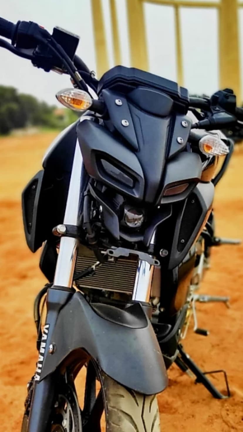 Yamaha MT-15 motorcycle with sharp headlights and front fender parked on sand with blurred green trees behind.