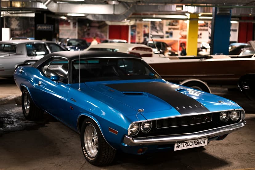 Blue Dodge Challenger with a black racing stripe and chrome accents displayed at an indoor vintage auto show.