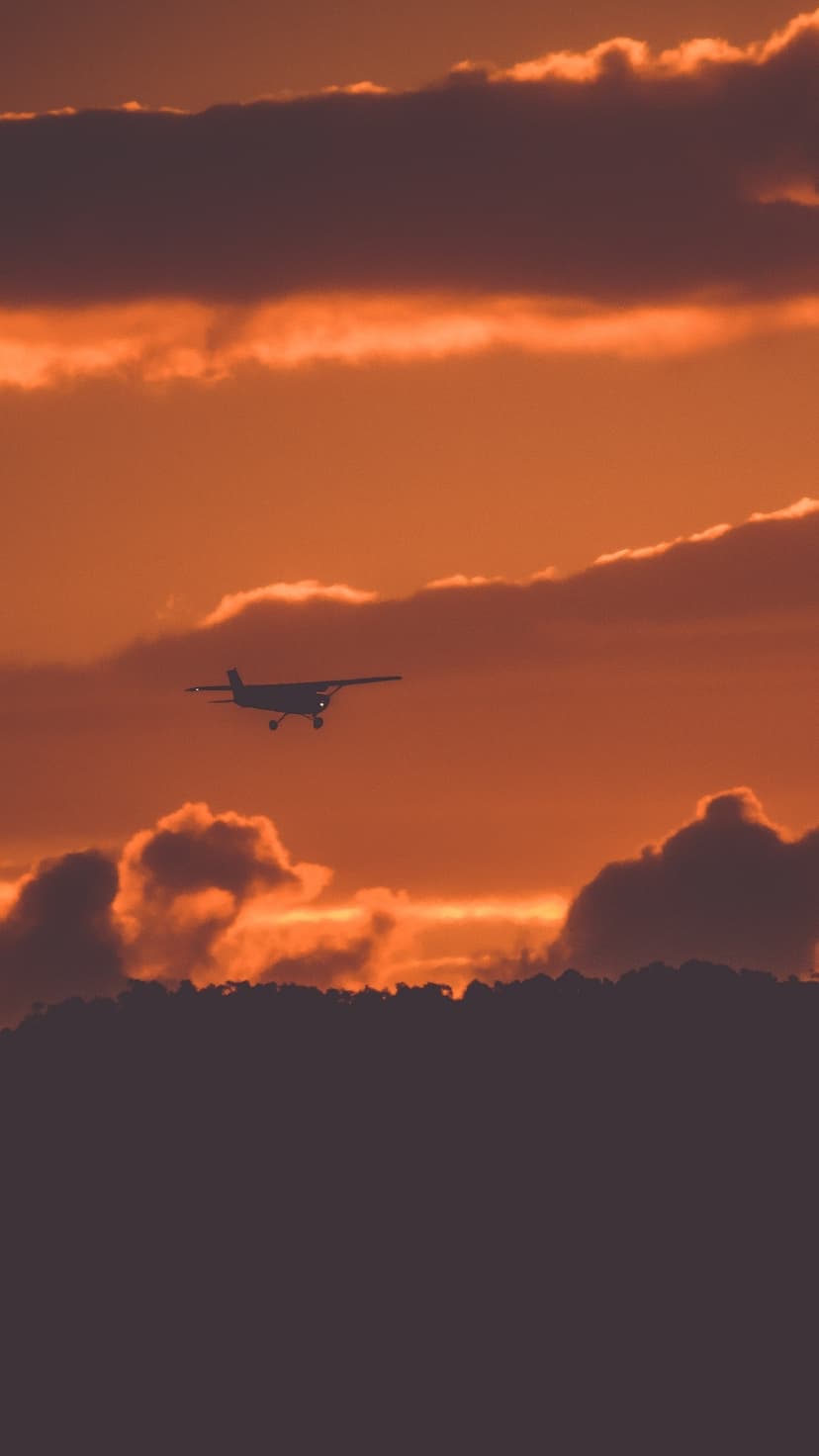 Airplane silhouette flying through fiery orange sunset clouds above a dark horizon with faint navigation lights.