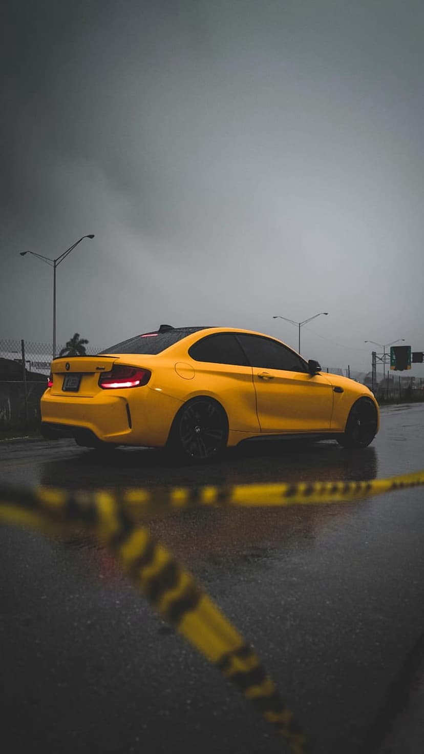 Yellow BMW M2 coupe parked on wet asphalt behind blurred caution tape under a dark, stormy sky with taillights lit.