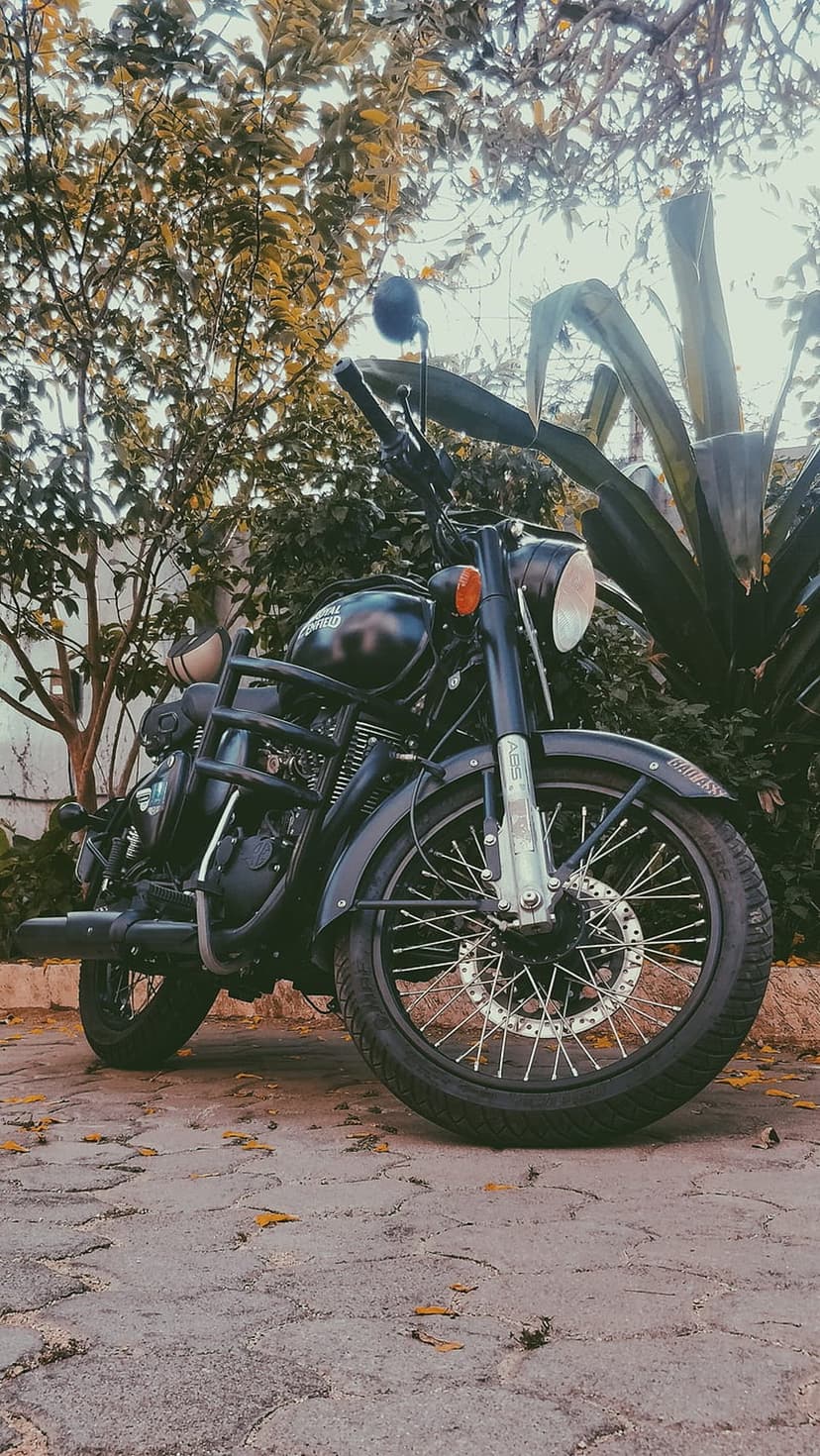 Vintage black motorcycle with chrome accents parked on a cobblestone path surrounded by green trees and sunlight.