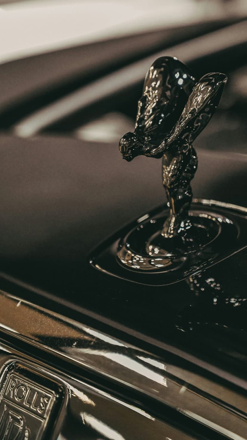Spirit of Ecstasy hood ornament rendered in polished metal stands on a reflective black Rolls-Royce car surface.