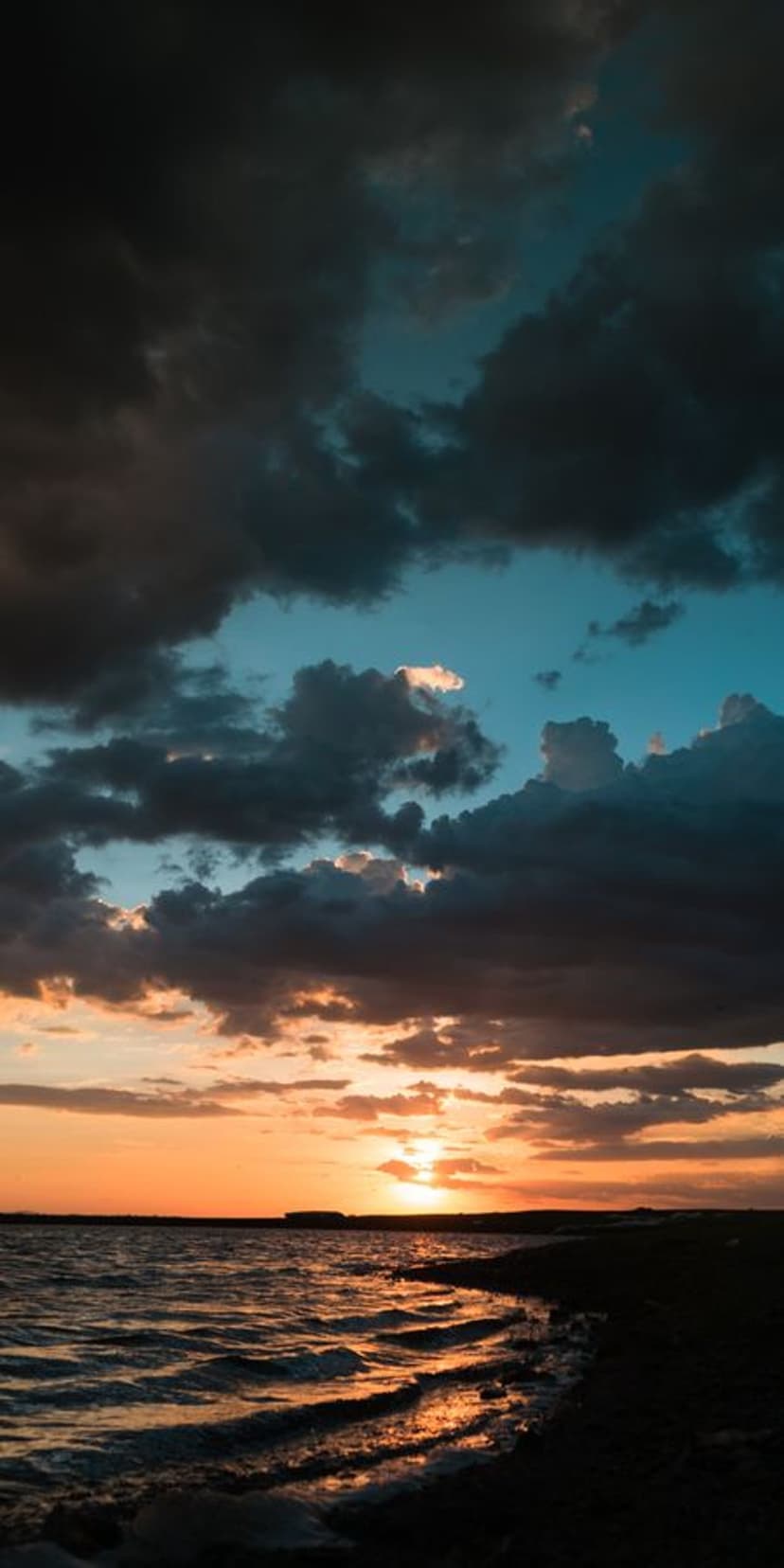 Fiery orange sunset over a dark ocean horizon with churning waves and heavy indigo clouds in a vertical frame.