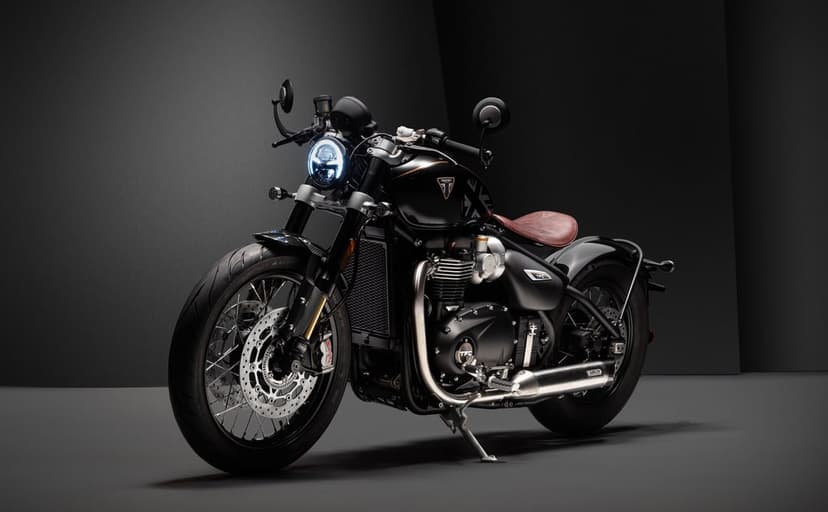 Black Triumph Bobber TFC motorcycle with a brown leather seat and glowing LED headlight in a dark gray studio.
