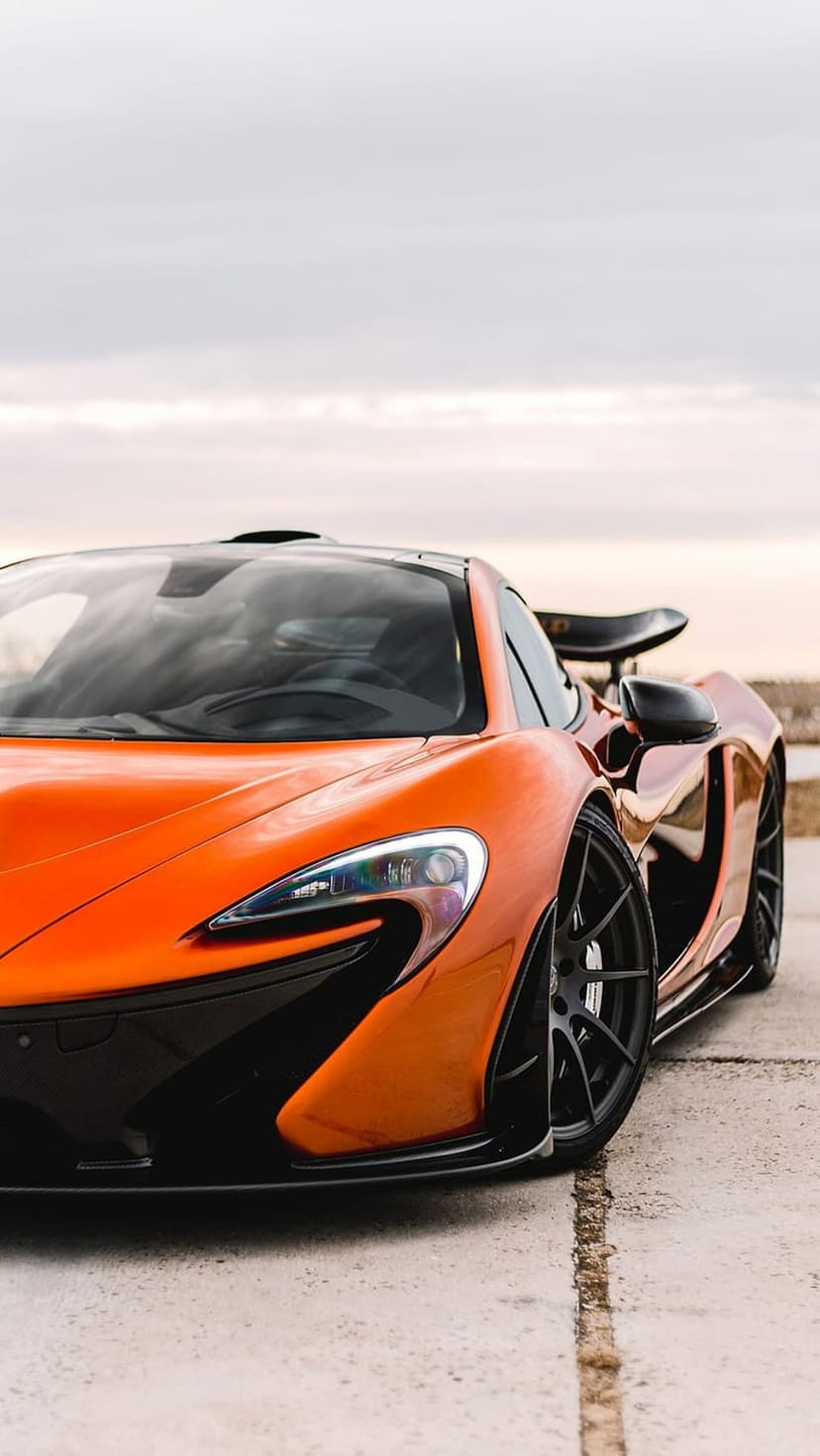 Orange McLaren P1 supercar with black carbon accents parked on gray concrete with a soft coastal horizon.