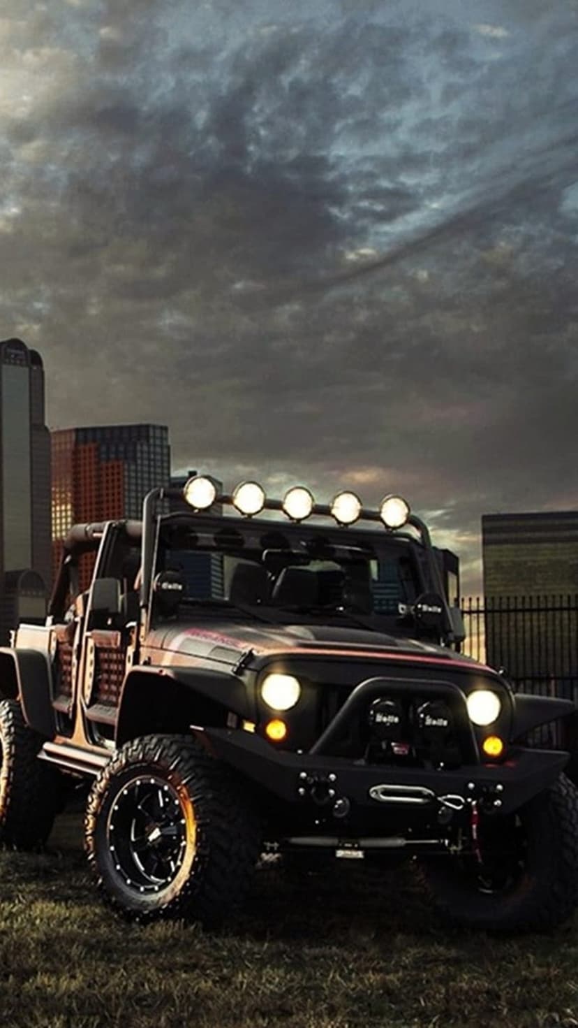 Black Jeep Wrangler with off-road tires and winch parked before a lit city skyline during twilight hours.