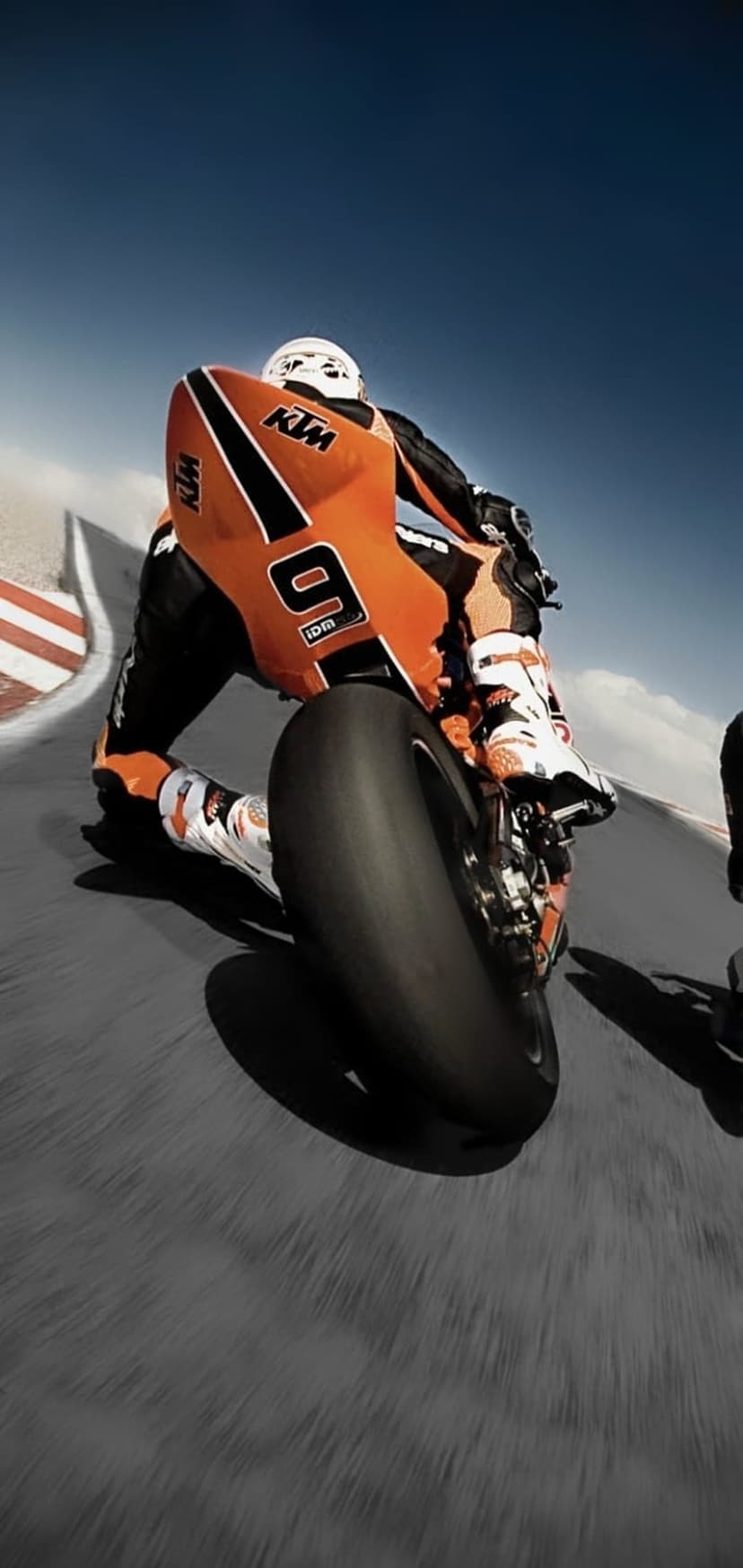 Motorcyclist in orange gear leans into a racetrack corner with a blue sky and blurred motion in the background.