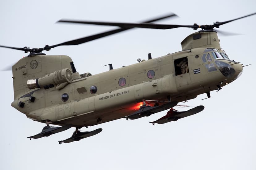 CH-47 Chinook helicopter in desert camouflage flies against a pale sky with twin rotors spinning and army markings.
