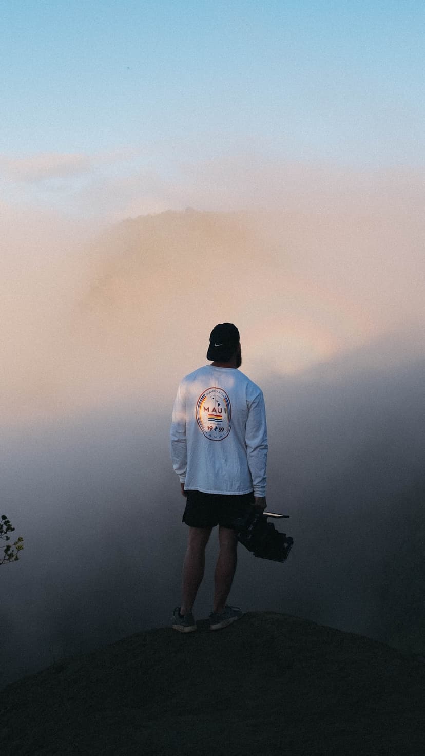 A person in a graphic shirt stands on a rocky peak overlooking thick white fog during a soft golden sunrise.