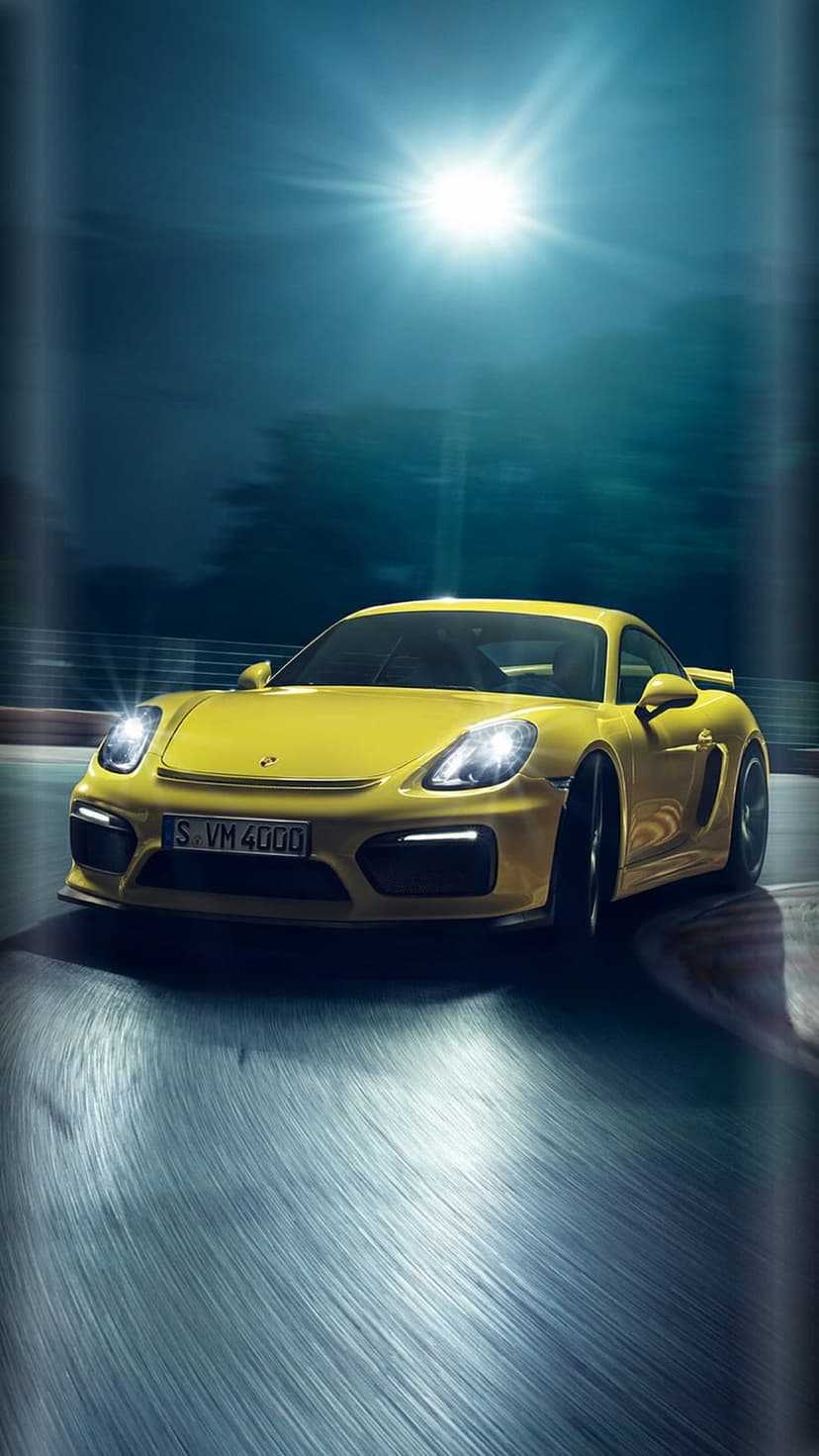Yellow Porsche Cayman GT4 on a dark wet racetrack illuminated by a bright overhead spotlight at night.