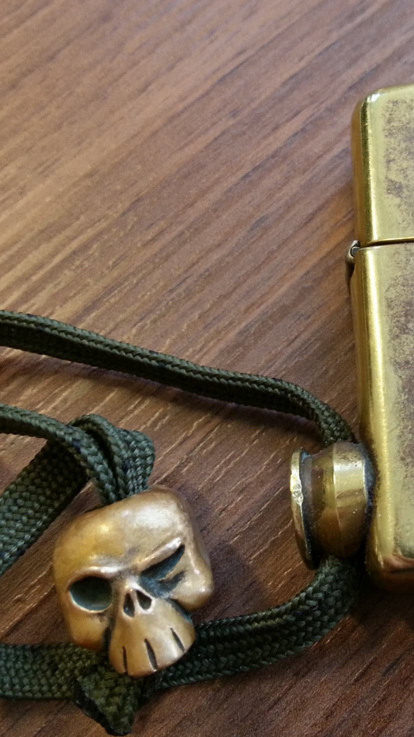 Brass skull bead and weathered metal lighter on a dark wood surface with a green braided paracord lanyard.