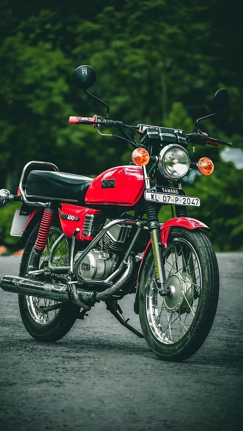 Red Yamaha RX 100 motorcycle with chrome accents parked on a road against a lush green forest background.