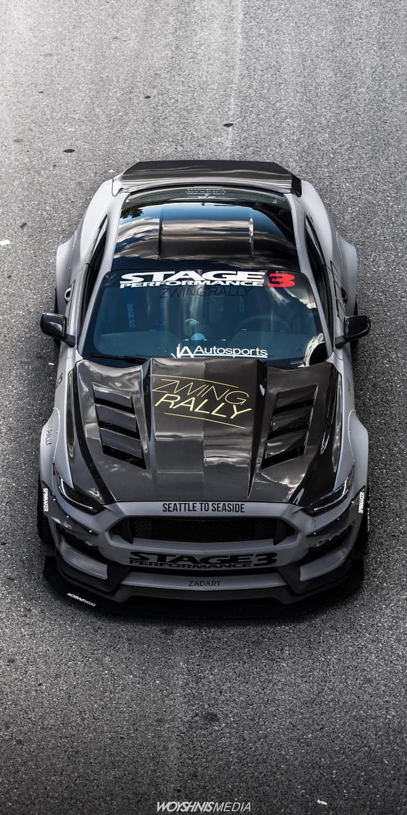 Grey widebody Ford Mustang with racing decals and carbon fiber hood seen from a top-down drone perspective.