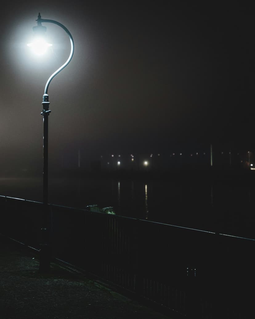 Ornate glowing lamp post stands beside a railing overlooking foggy water with distant blurred lights at night.