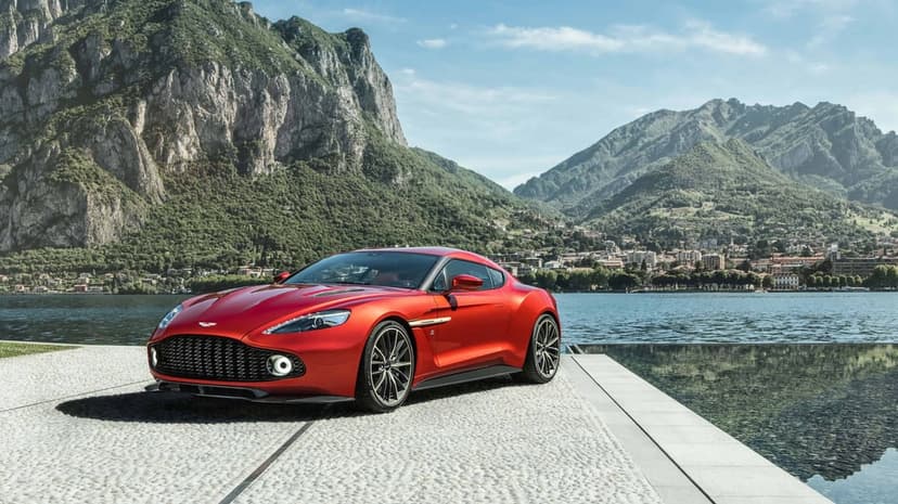 Red luxury sports car parked on a stone pier against a backdrop of blue lake water and towering green mountains.