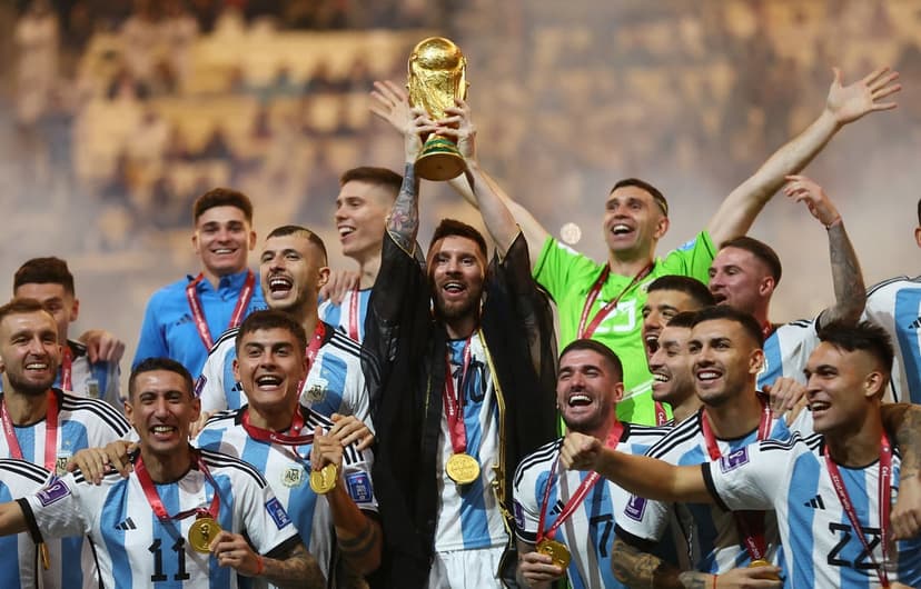 Lionel Messi hoists the gold FIFA World Cup trophy while surrounded by teammates in blue and white jerseys.