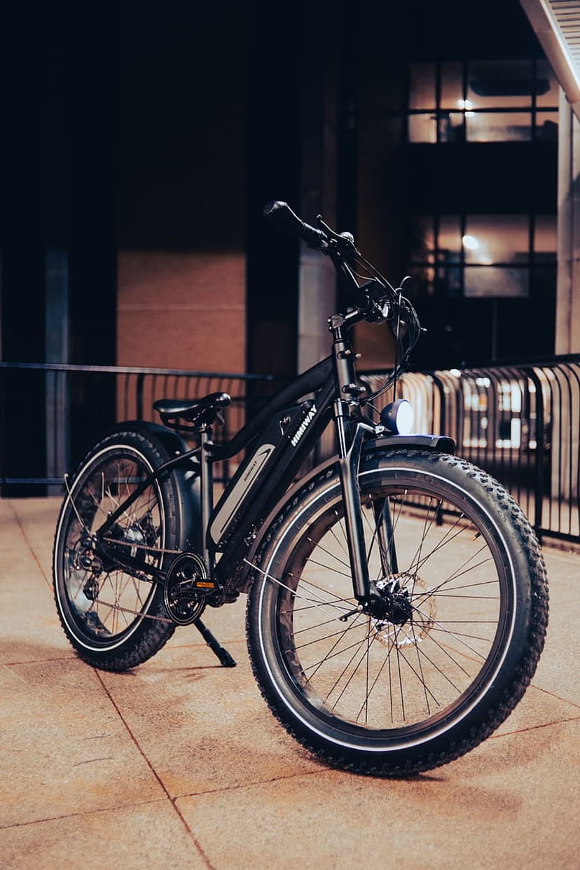 Black fat tire electric bike with a front headlight parked on a stone walkway against blurred city lights at night.