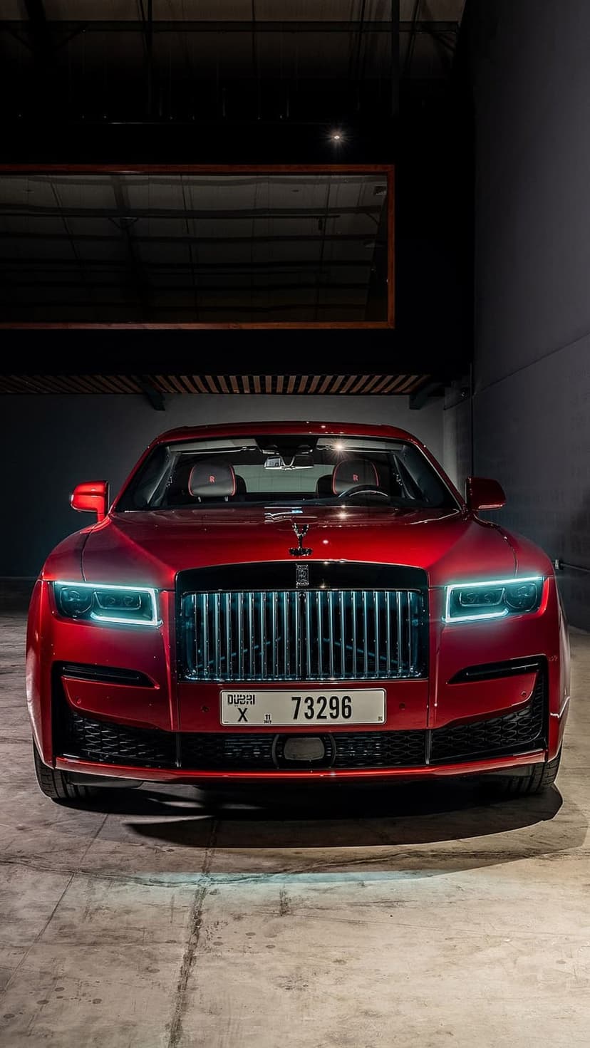 Red Rolls-Royce Ghost parked in a dark minimalist garage with glowing headlights and a chrome grille.