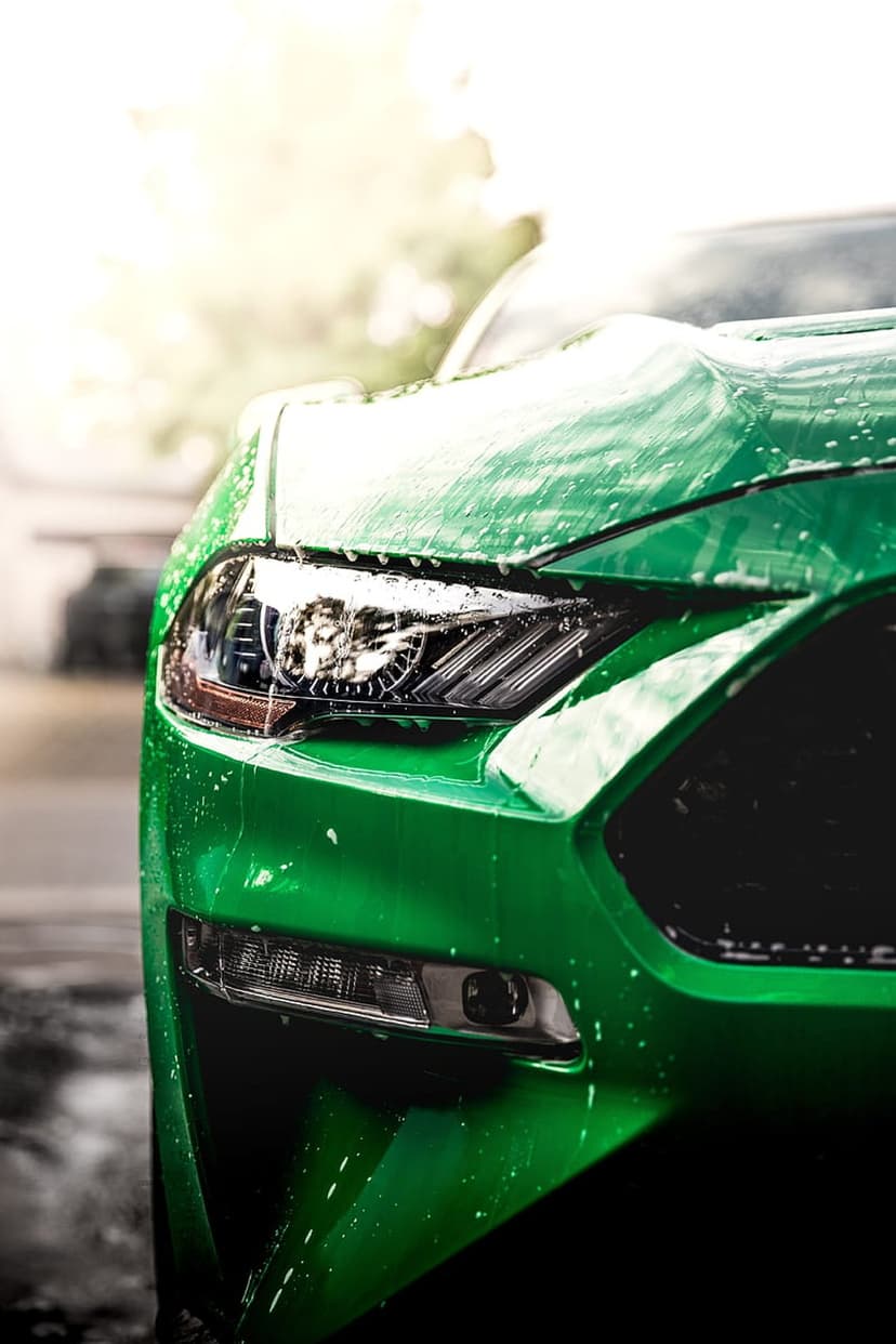 Green Ford Mustang front headlight and grille covered in soap foam and water droplets under bright sunlight.