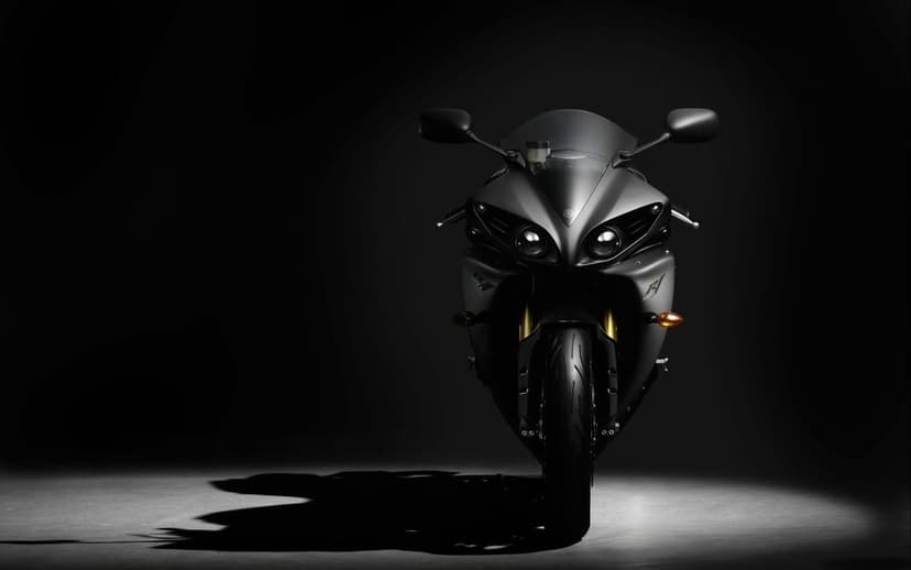 Front view of a matte black Yamaha R1 motorcycle with golden forks and lit headlights in a dark studio.