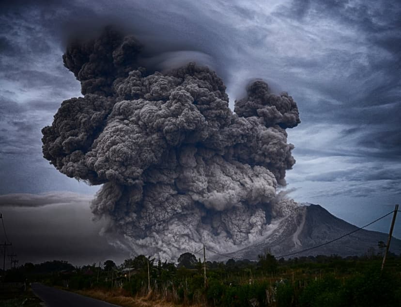 Massive gray ash cloud billowing from a cone-shaped volcano under a dark stormy sky with a rural road below.