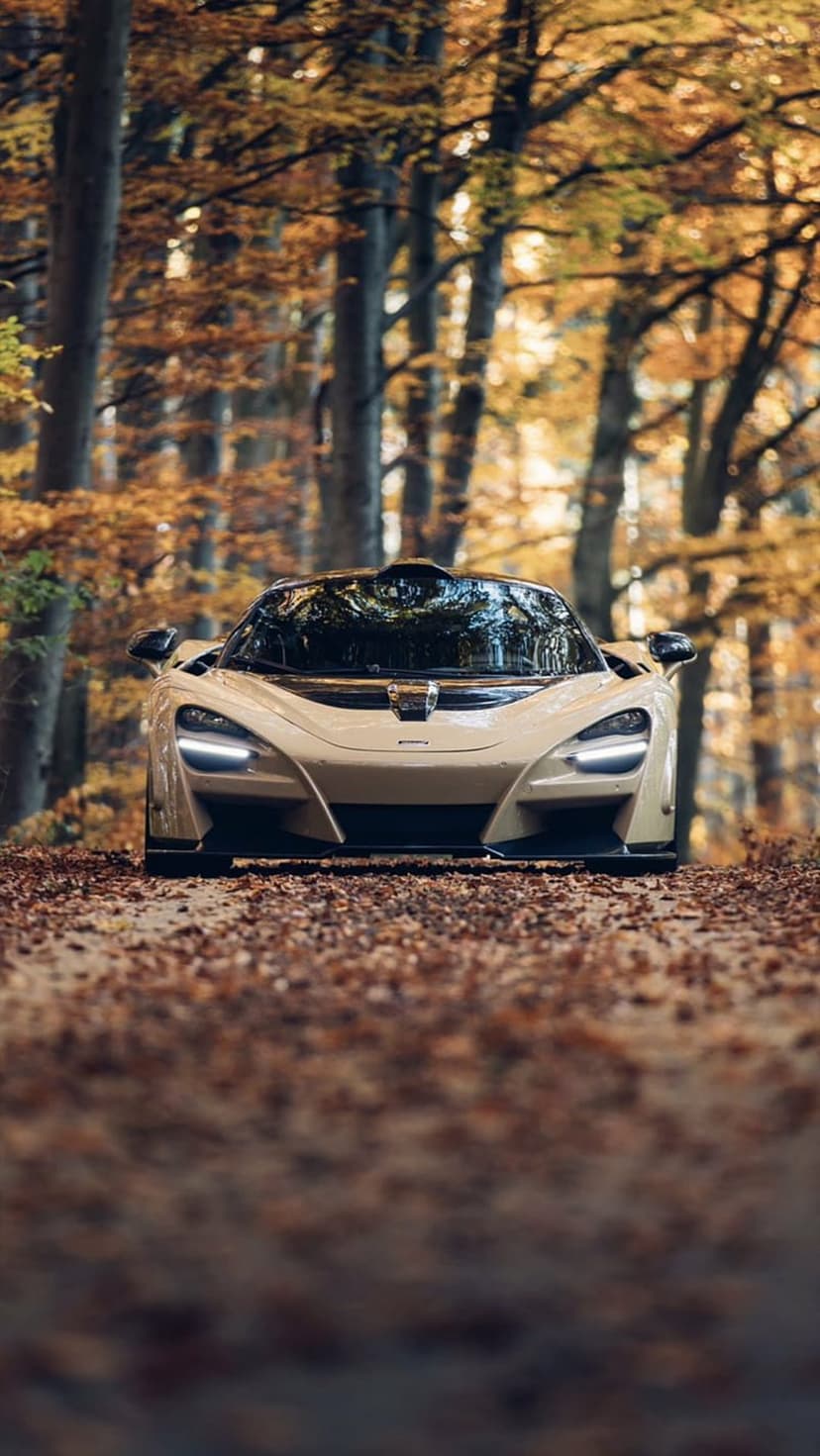 McLaren 720S in beige parked on an asphalt road covered in orange autumn leaves within a dense forest of tall trees.