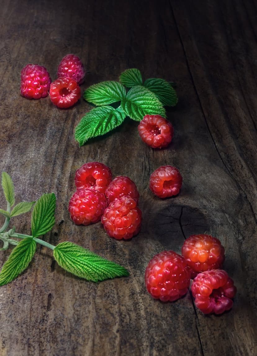 Ripe red raspberries and fresh green leaves scattered over a dark, textured wooden surface with soft natural light.