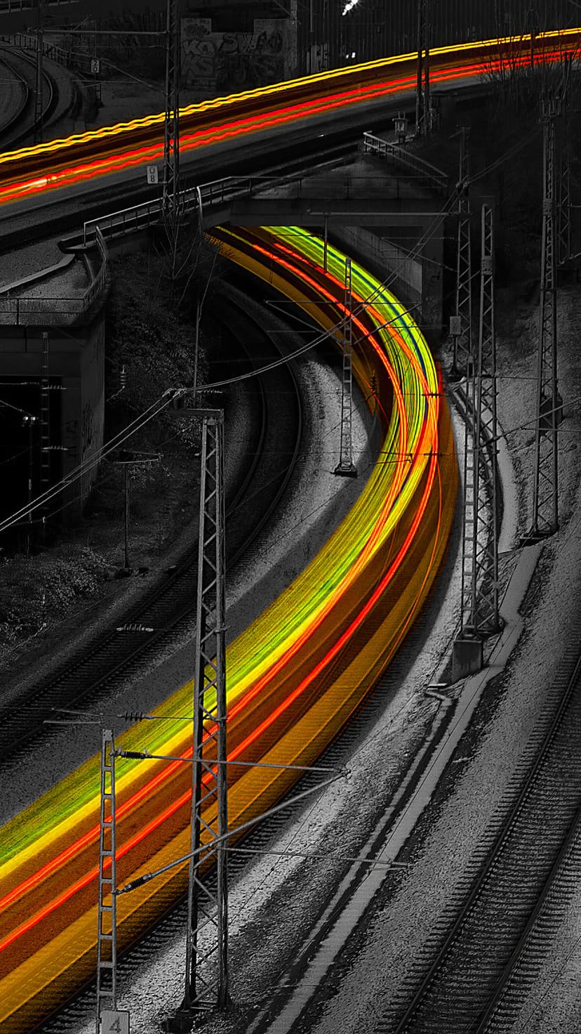 Red and yellow train light trails curve through a black and white industrial landscape with power lines.