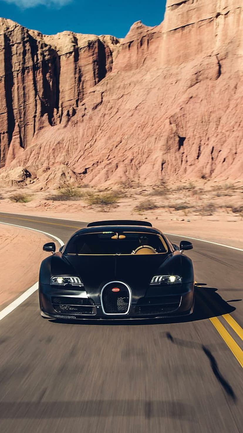 Black Bugatti Veyron supercar driving on a winding desert road surrounded by massive red rock canyon walls.