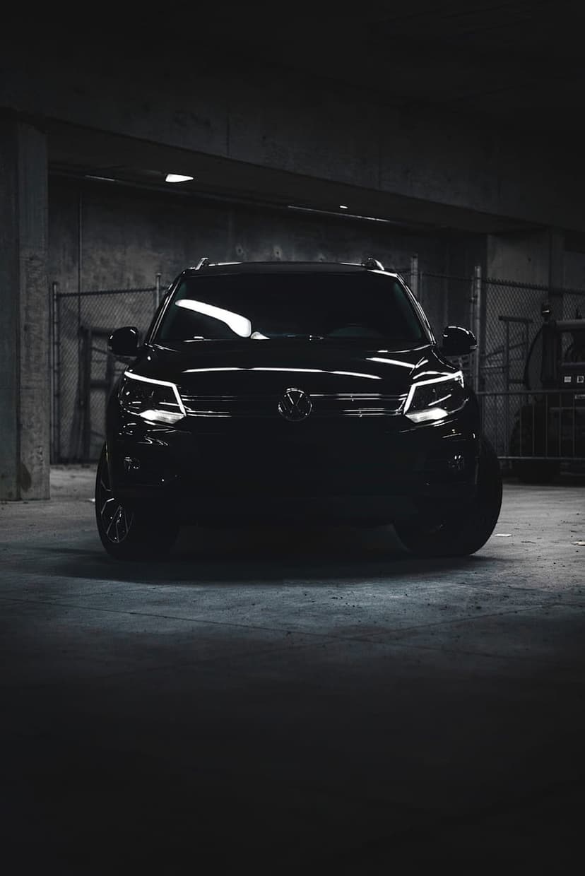 Black Volkswagen Tiguan SUV with glowing headlights parked against a chain-link fence in a dark garage.