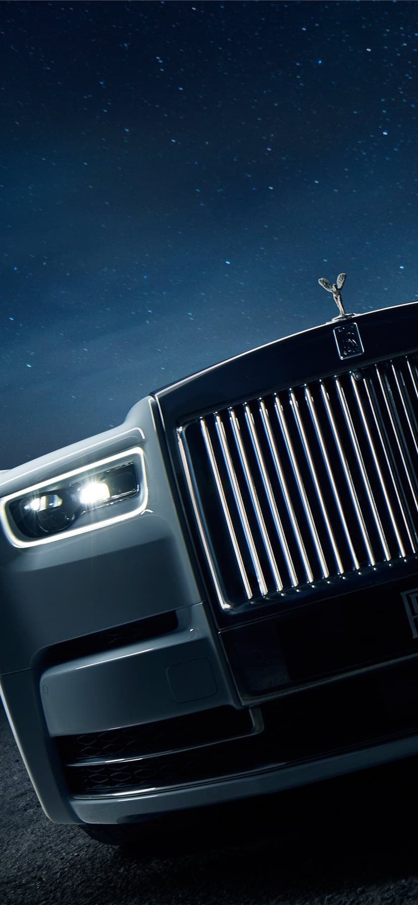 Luxury car front grille and glowing LED headlights under a dark blue night sky filled with bright stars.