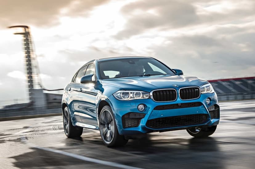 Blue BMW X6 M speeding on a wet racetrack with a blurred tower and cloudy sky background.