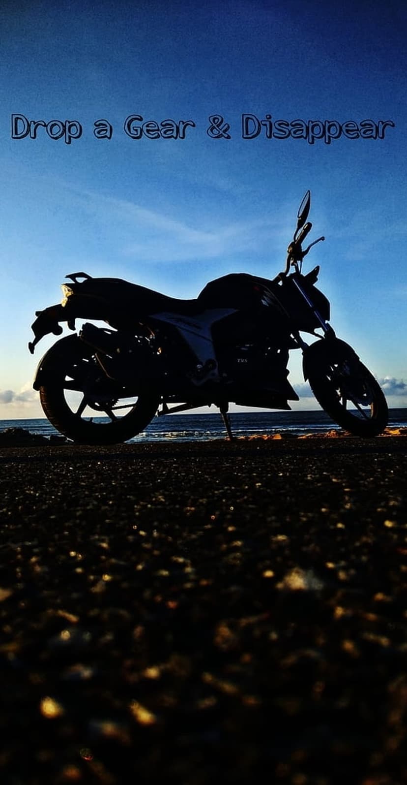 Black motorcycle silhouette on a pebble beach with a vibrant blue sunset sky and coastal waves in the distance.