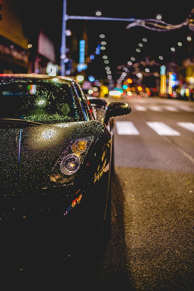 Black sports car with raindrops on its hood parked on a wet city street at night under glowing streetlights.