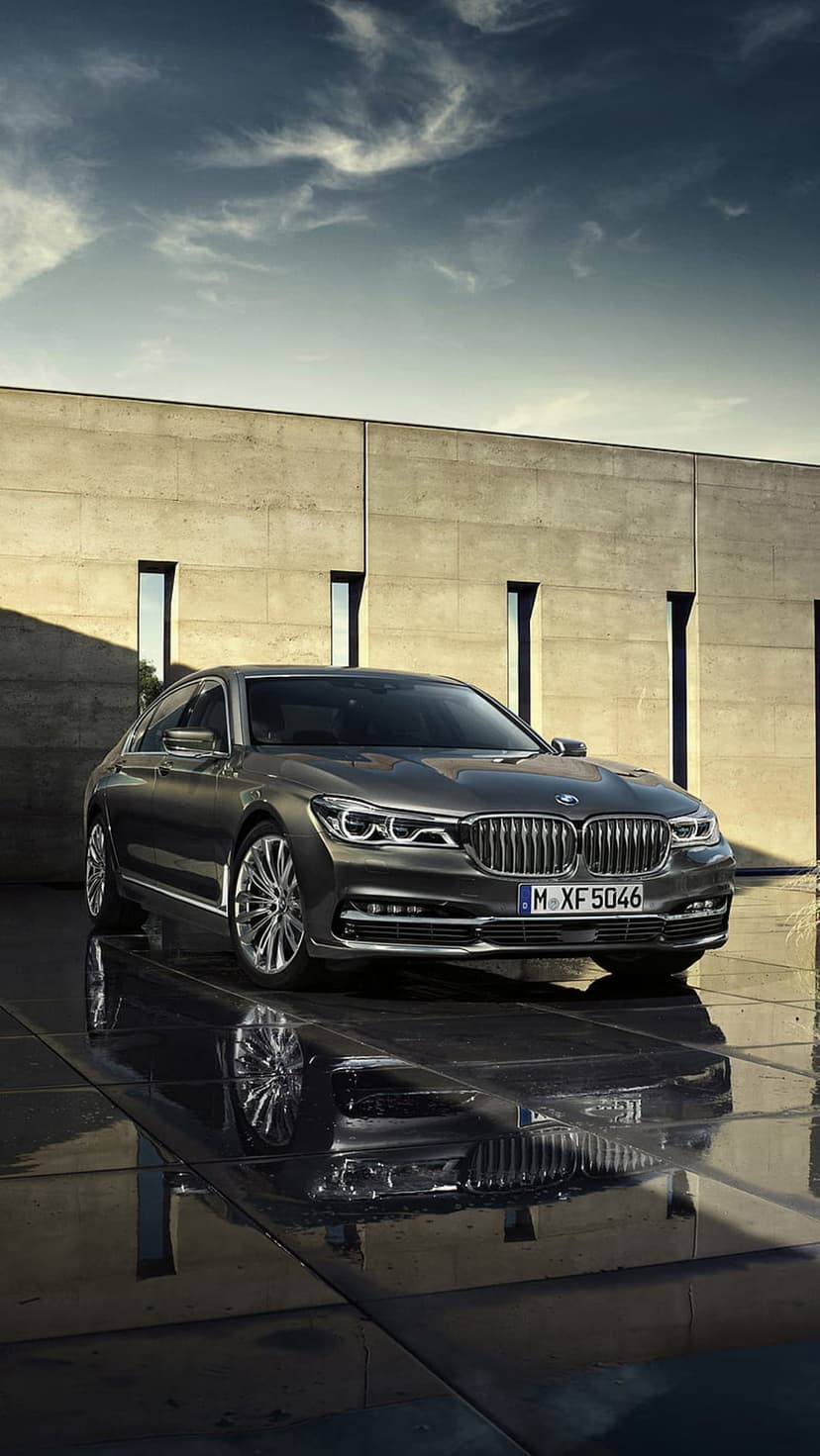 Dark grey BMW sedan parked on a wet, reflective urban street beside a modern concrete building under a cloudy sky.