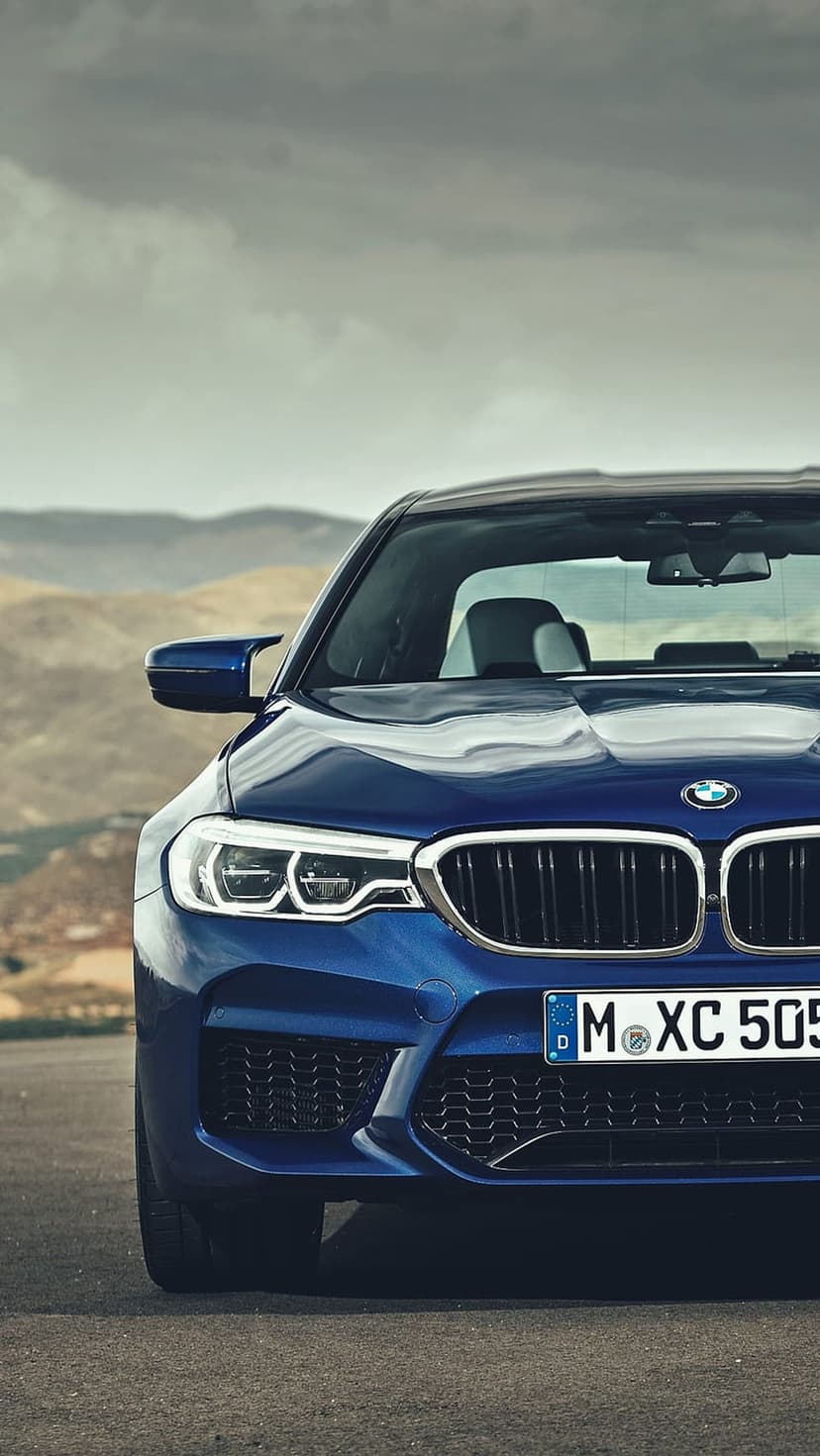 Blue BMW M5 sedan parked on an asphalt mountain road with rolling green hills and a cloudy sky in the background.