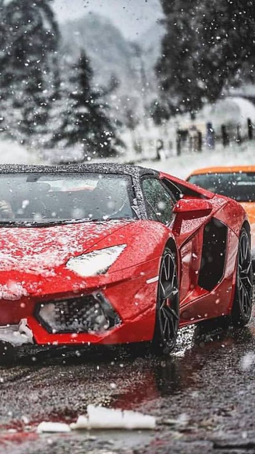 Red Lamborghini supercar on a snowy asphalt road with falling snow, winter trees, and a distant orange car.