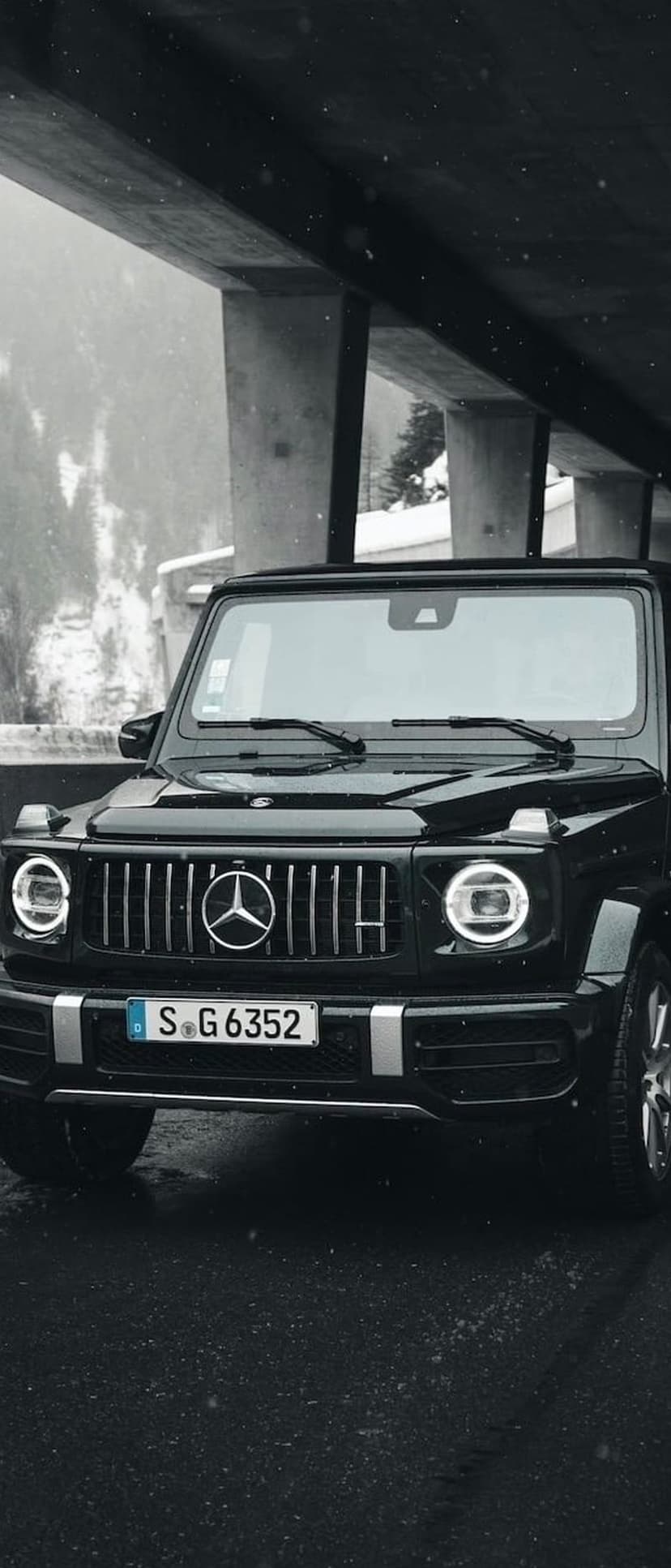 Black Mercedes G-Wagon SUV parked beneath a concrete bridge during a heavy snowfall on wet asphalt at night.
