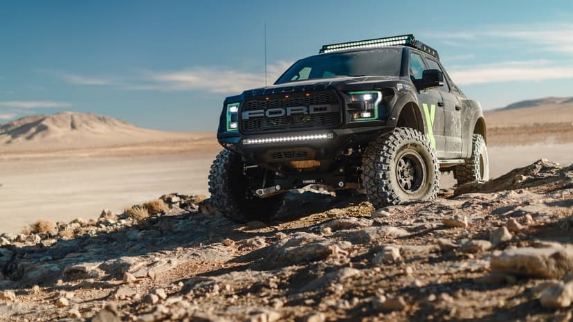 Black Ford Raptor truck with green headlights on rocky desert ground under a bright clear blue sky.