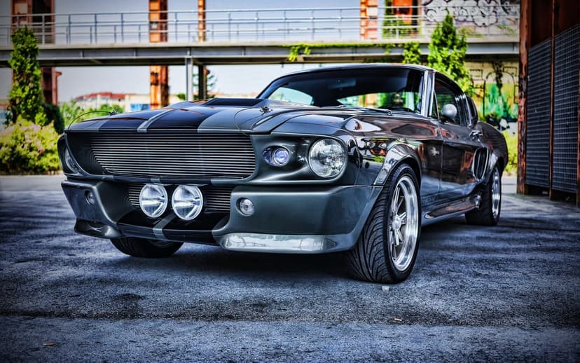 Classic gray muscle car with black racing stripes and chrome wheels parked in an industrial urban setting.